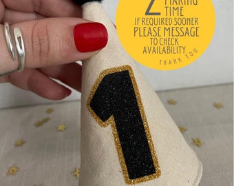 May include: A white fabric party hat with a black felt ball on top and a gold glitter outlined black number one on the front. The hat is held by a hand. A yellow circle with black text says "2 week making time if required sooner please message to check availability thank you".