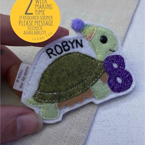 May include: A felt turtle with the name "ROBYN" embroidered on it. The turtle has a green shell, light green head and feet, and a purple glitter number "8". The image also includes text that says "2 WEEK MAKING TIME".