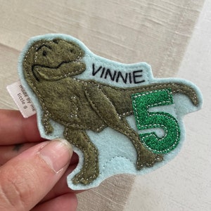 May include: A light blue felt patch with a green felt number 5 and a brown felt dinosaur. The dinosaur is facing right and the number is to the right of the dinosaur. The patch has the name "Vinnie" stitched in black thread.