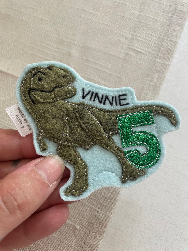 May include: A light blue felt patch with a green felt number 5 and a brown felt dinosaur. The dinosaur is facing right and the number is to the right of the dinosaur. The patch has the name "Vinnie" stitched in black thread.