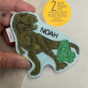 May include: A light blue felt dinosaur applique with green glitter accents. The dinosaur is holding a green glitter number 4. The applique is personalised with the name "NOAH".