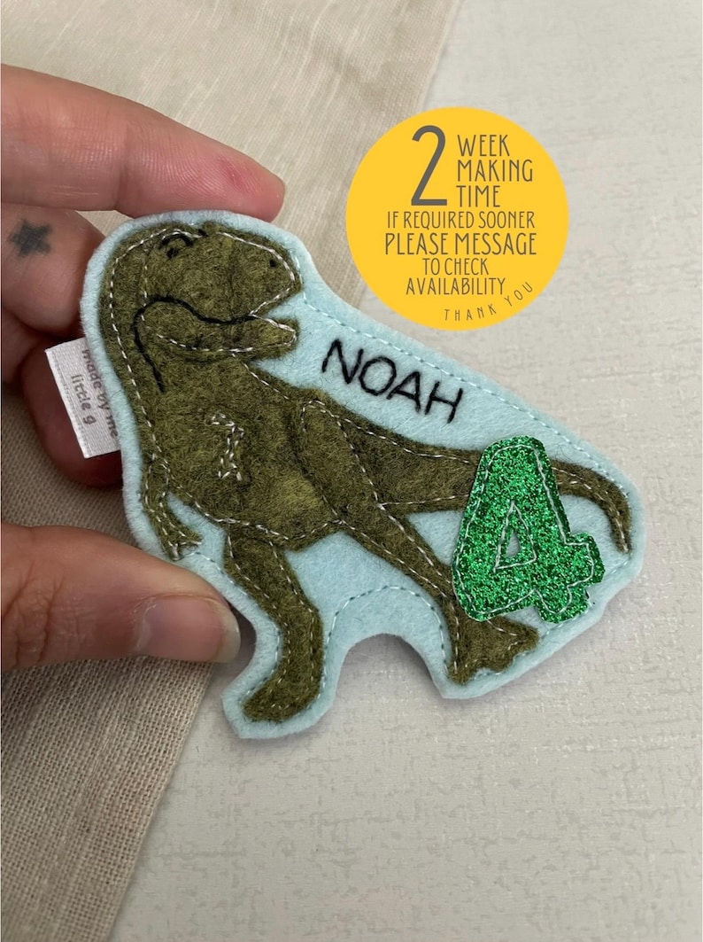 May include: A light blue felt dinosaur applique with green glitter accents. The dinosaur is holding a green glitter number 4. The applique is personalised with the name "NOAH".