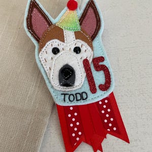 May include: A handmade felt dog-themed birthday badge. The badge features a dog's face with a party hat, the number 15 in glitter, and the name "TODD". Red ribbon with white polka dots completes the design.