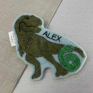 May include: Green felt dinosaur applique with the name "ALEX" and a green glitter number "6" on a light blue background. The applique is made by "little g" and is perfect for a birthday party or craft project.