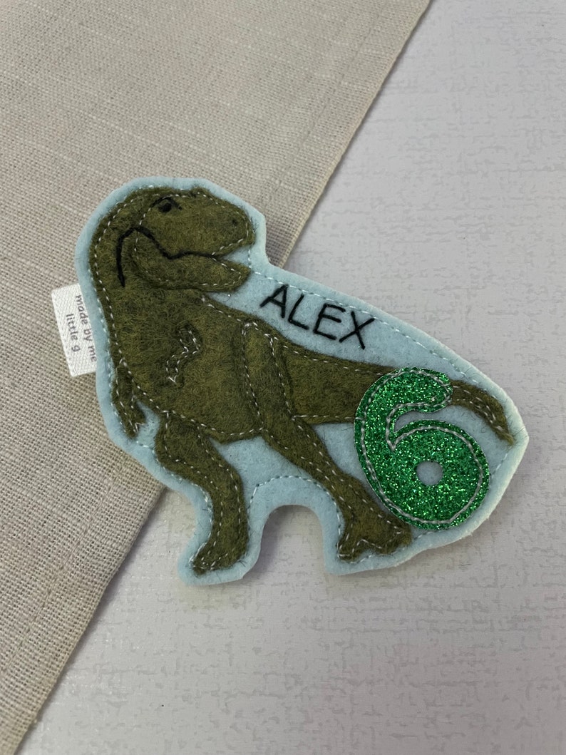 May include: Green felt dinosaur applique with the name "ALEX" and a green glitter number "6" on a light blue background. The applique is made by "little g" and is perfect for a birthday party or craft project.