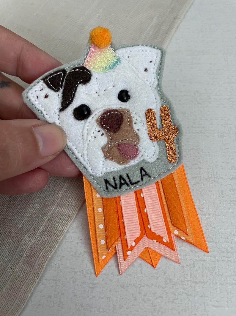 May include: A handmade felt dog birthday badge with the name "NALA" and the number "4". The dog's head is white with brown spots and a party hat. Orange and pink ribbons hang below the badge.