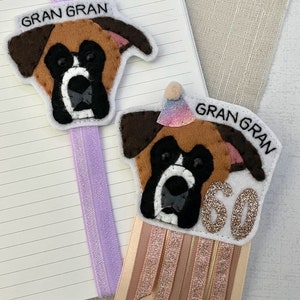 May include: Two felt dog-shaped bookmarks. One is attached to a purple ribbon. The other has a party hat and a ribbon with the number 60. Both have the text "GRAN GRAN". The dogs are brown, black, and white.