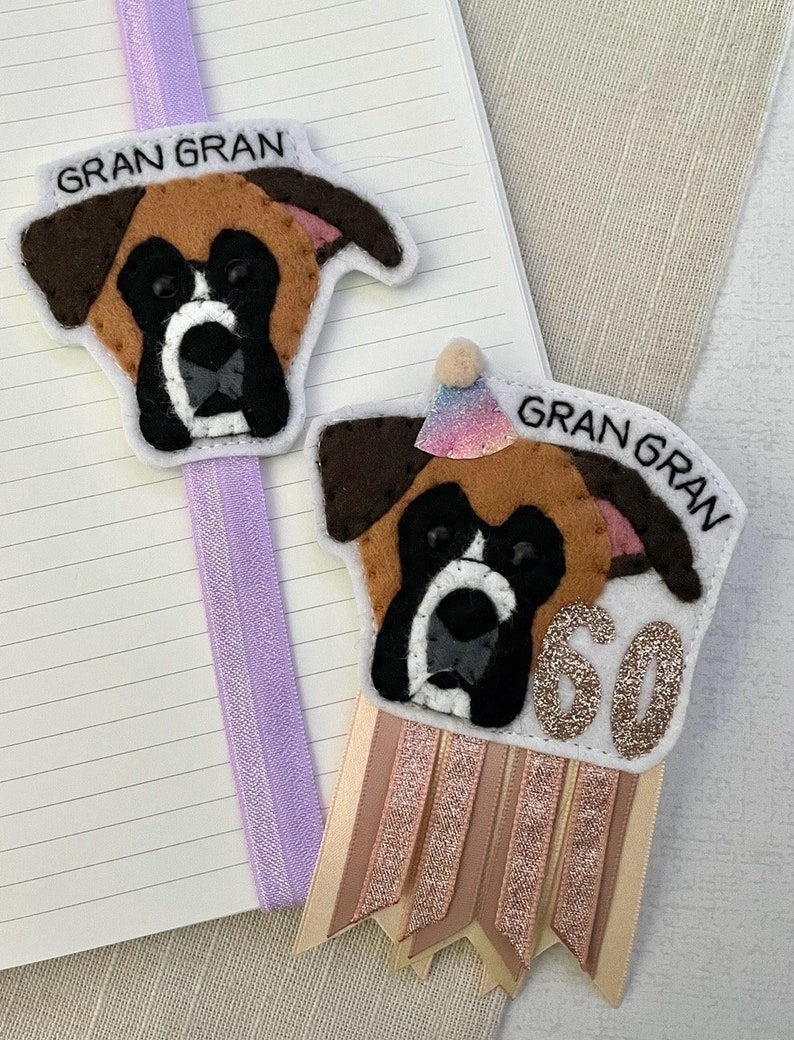 May include: Two felt dog-shaped bookmarks. One is attached to a purple ribbon. The other has a party hat and a ribbon with the number 60. Both have the text "GRAN GRAN". The dogs are brown, black, and white.