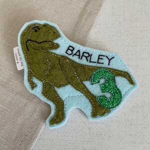 May include: A light blue felt dinosaur applique with green glitter accents. The dinosaur is a T-Rex with a stitched outline. The applique says "BARLEY" and "3" in black text.