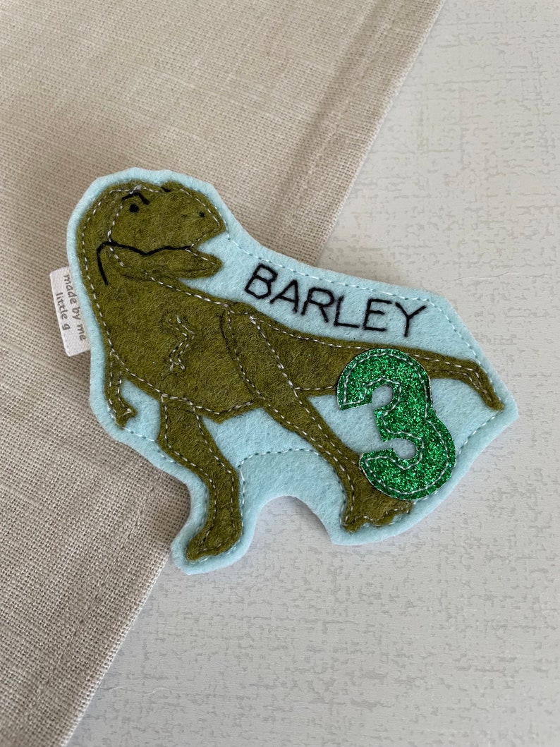 May include: A light blue felt dinosaur applique with green glitter accents. The dinosaur is a T-Rex with a stitched outline. The applique says "BARLEY" and "3" in black text.