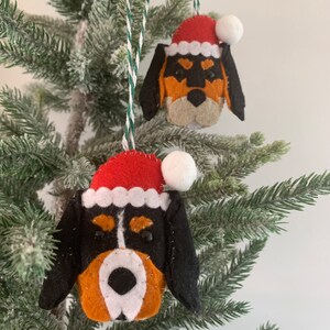 May include: Two felt Christmas ornaments shaped like dog heads wearing Santa hats. The ornaments are black, white, and orange.
