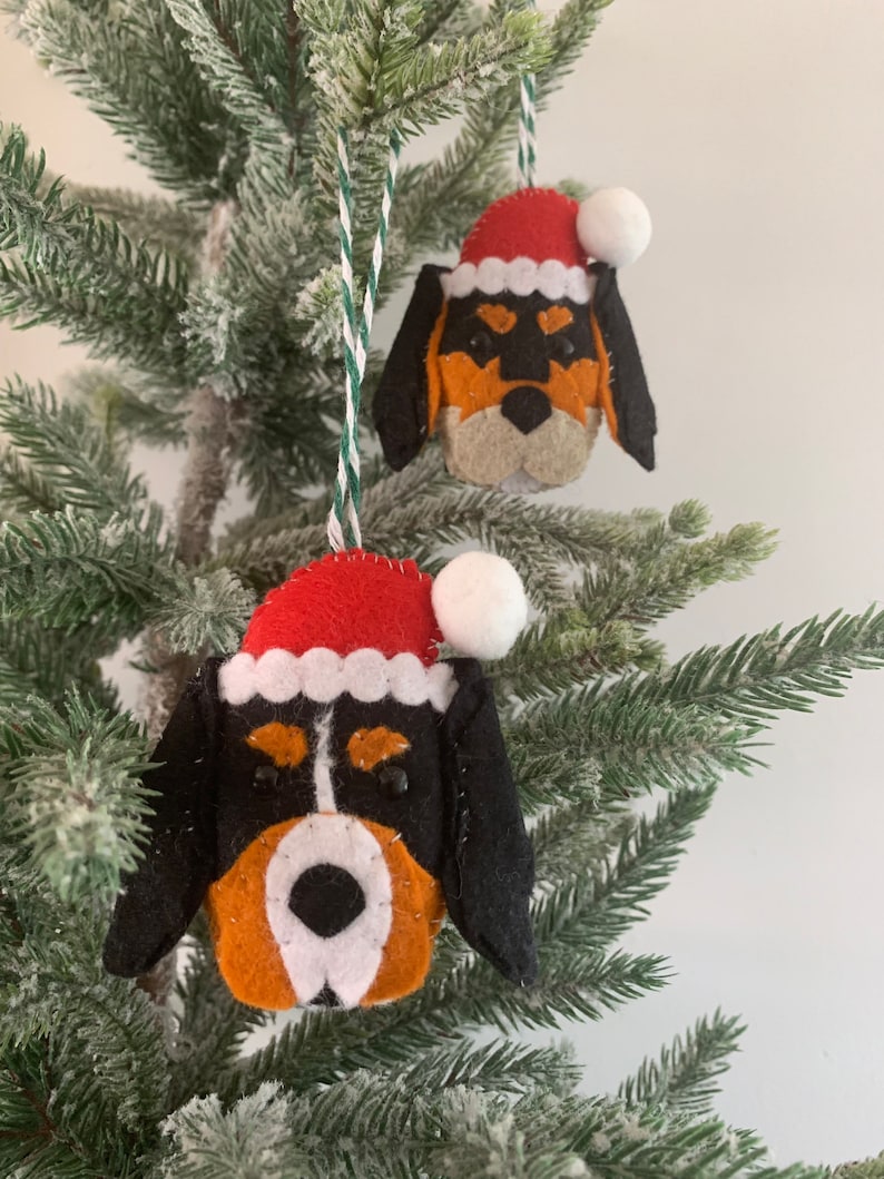May include: Two felt Christmas ornaments shaped like dog heads wearing Santa hats. The ornaments are black, white, and orange.