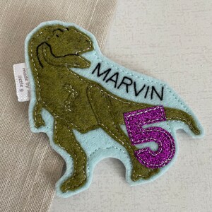 May include: A light blue felt dinosaur applique with green felt detailing. The dinosaur is standing on its hind legs with its mouth open. The dinosaur has the name "MARVIN" embroidered on its side and a purple felt number "5" on its tail.