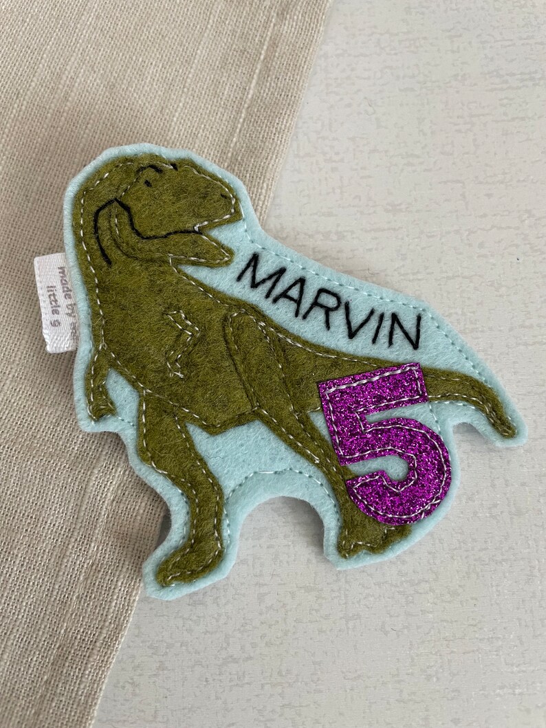 May include: A light blue felt dinosaur applique with green felt detailing. The dinosaur is standing on its hind legs with its mouth open. The dinosaur has the name "MARVIN" embroidered on its side and a purple felt number "5" on its tail.