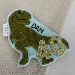 May include: A green felt dinosaur with a blue felt background. The dinosaur has a name tag that says "DAN" and a gold glitter number "40" on the background. The tag says "made by me little g".
