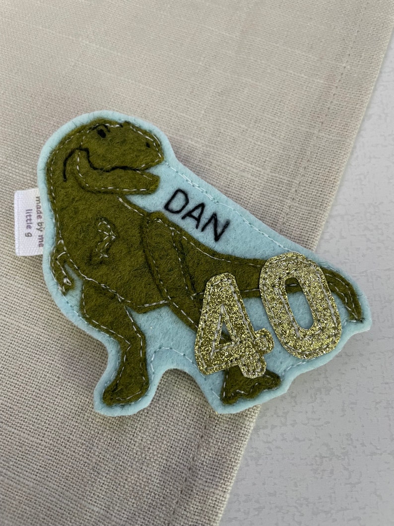 May include: A green felt dinosaur with a blue felt background. The dinosaur has a name tag that says "DAN" and a gold glitter number "40" on the background. The tag says "made by me little g".