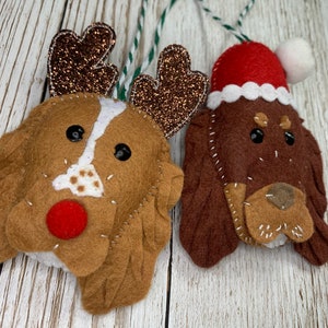 May include: Two felt Christmas ornaments shaped like dogs. One dog is wearing a Santa hat and the other has reindeer antlers. Both dogs have brown fur and black eyes.