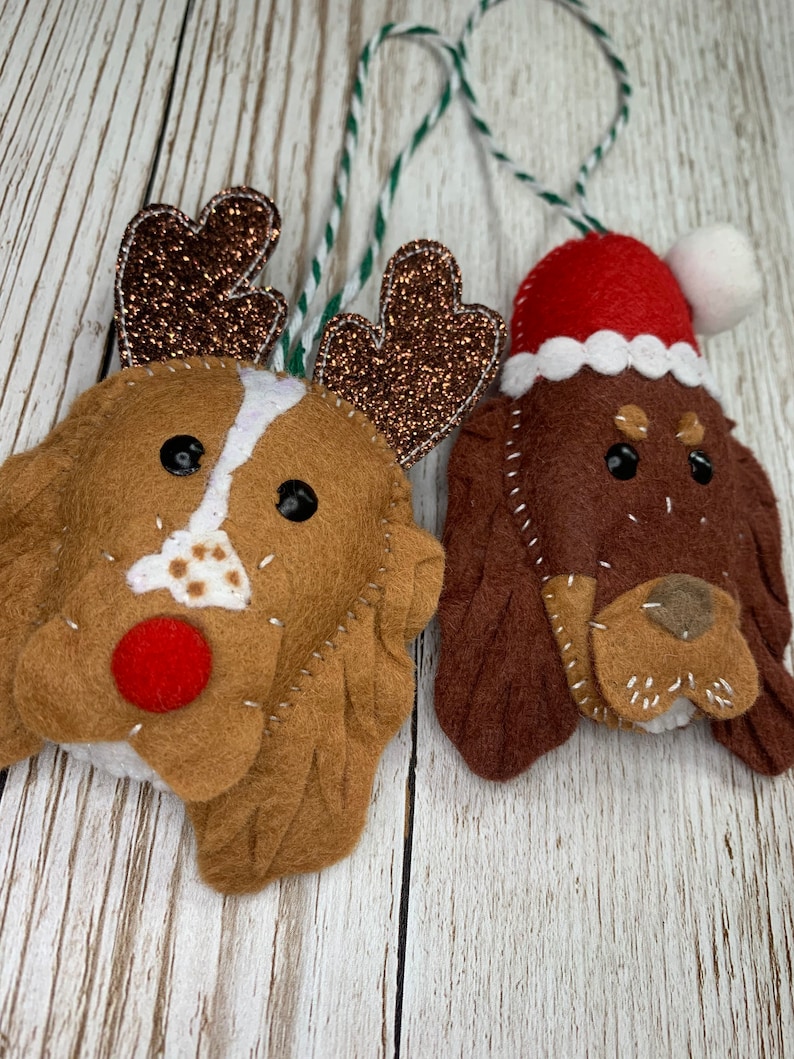 May include: Two felt Christmas ornaments shaped like dogs. One dog is wearing a Santa hat and the other has reindeer antlers. Both dogs have brown fur and black eyes.