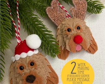 May include: Two felt Christmas ornaments shaped like a dog. One dog is wearing a Santa hat and the other has reindeer antlers. The ornaments are hanging from a Christmas tree branch. The text on the yellow circle reads: "2 week making time. If required sooner please message to check availability. Thank you."
