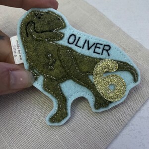 May include: Green felt dinosaur applique with a light blue felt background. The dinosaur has a name, "OLIVER", stitched in black thread and a gold glitter number "6" on the side.