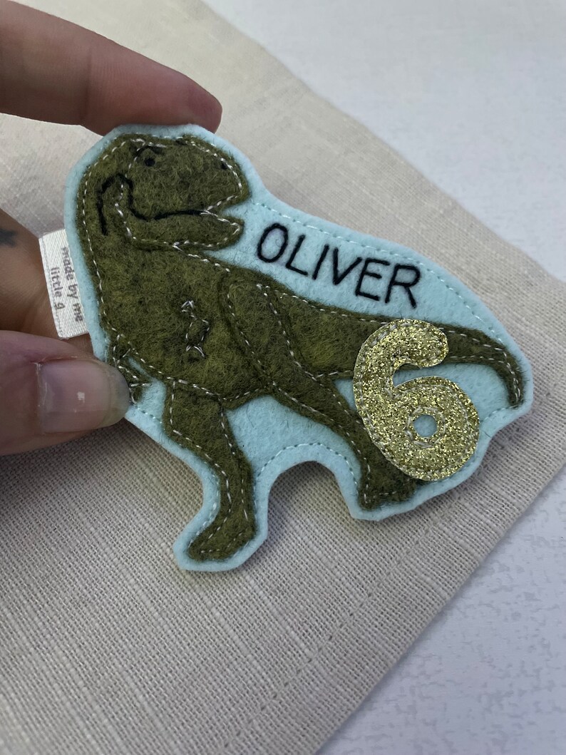 May include: Green felt dinosaur applique with a light blue felt background. The dinosaur has a name, "OLIVER", stitched in black thread and a gold glitter number "6" on the side.