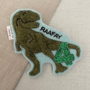 May include: A green felt dinosaur applique with a light blue felt background. The dinosaur is holding a green glitter number 4. The text "RAAFAY" is embroidered on the background. The text "made by me little g" is on a white tag attached to the dinosaur.