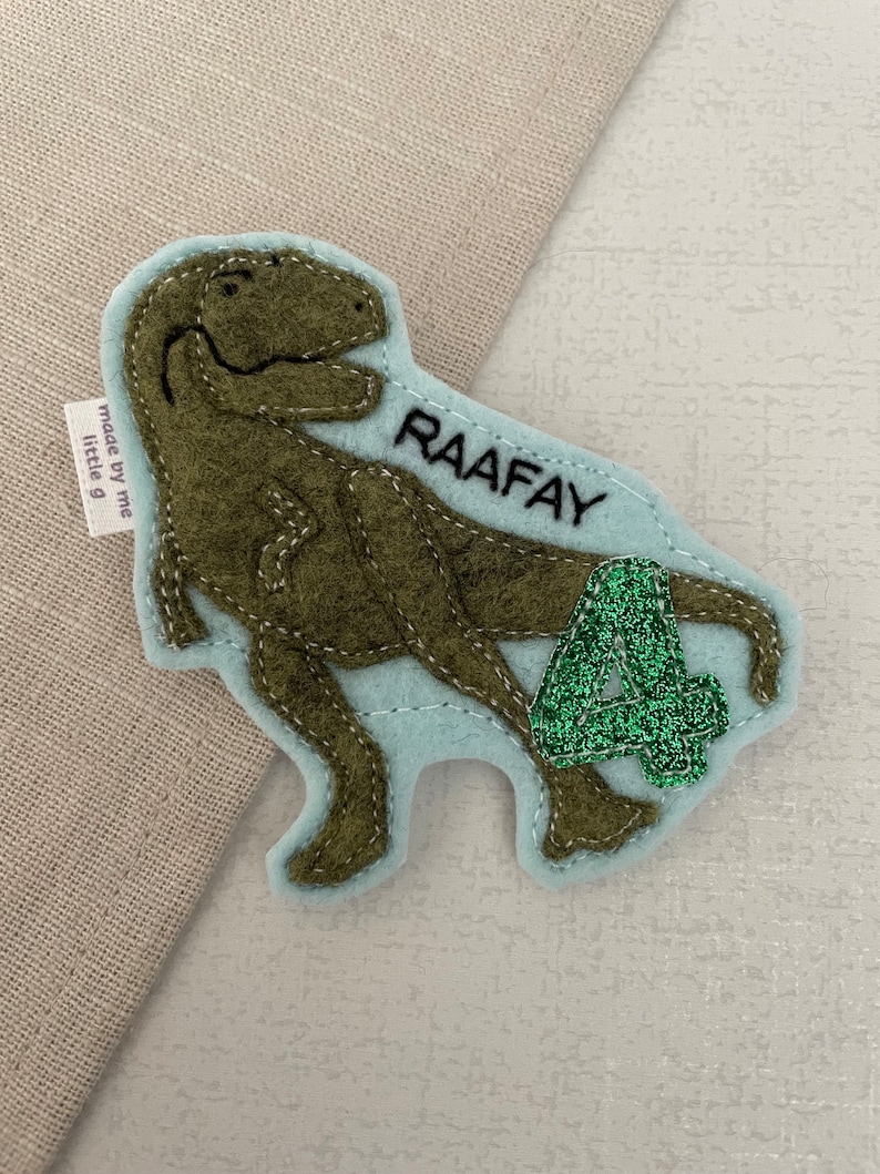 May include: A green felt dinosaur applique with a light blue felt background. The dinosaur is holding a green glitter number 4. The text "RAAFAY" is embroidered on the background. The text "made by me little g" is on a white tag attached to the dinosaur.