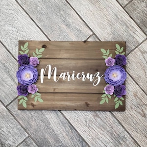 May include: Wooden sign with the name "Maricruz" written in white cursive lettering. The sign is decorated with purple paper flowers and green leaves.