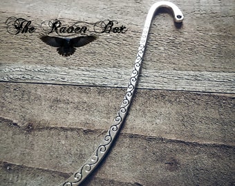 May include: Silver bookmark with a decorative scroll design. The bookmark is shaped like a long, thin rectangle with a curved top.