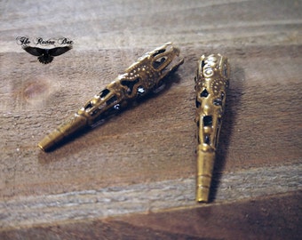 Puede incluir: Dos hallazgos de joyería de metal de color latón antiguo con intrincados diseños de filigrana. Los hallazgos son alargados, con forma cónica y con recortes decorativos. El logotipo de The Raven Box está en la esquina superior izquierda.