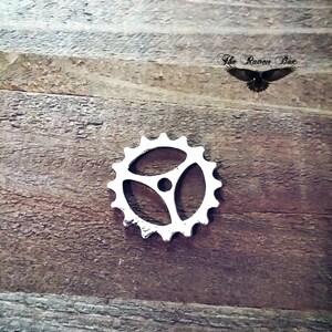 Op de afbeelding: Een zilveren metalen tandwiel met een driepuntige stervorm. Het tandwiel ligt op een houten oppervlak.