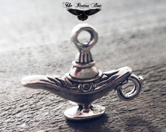 Puede incluir: Un amuleto de plata con forma de lámpara de genio adornada. La lámpara tiene un bucle en la parte superior para sujetarlo a un collar o pulsera.