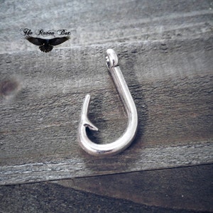 Pode incluir: Encanto de anzol de pesca de metal prateado. O anzol mede aproximadamente 5 cm de comprimento e tem uma ponta afiada.