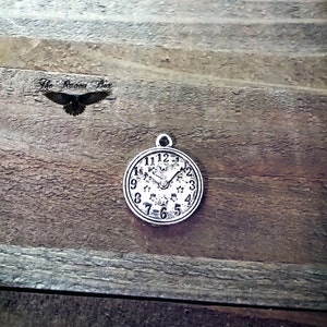 May include: Silver charm in the shape of a clock face with black Roman numeral markings and a star design. The clock hands are set to 10:50.