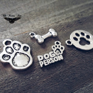 May include: Five silver charms with dog-themed designs. The charms include a paw print, a bone, a paw print inside a circle, a paw print, and the words "Dog Person".