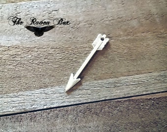 May include: Silver arrow charm with a pointed tip and a fletched tail. The arrow is on a wooden surface.