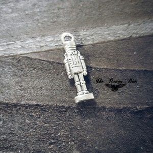 May include: Silver-toned metal charm shaped like a nutcracker soldier. The charm has a detailed design with a hat, a mustache, and a sword.