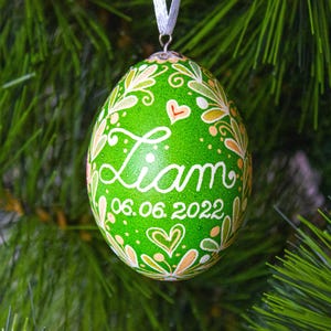 Pode incluir: Um enfeite de Natal verde em forma de ovo com o nome "Liam" e a data "06.06.2022" em escrita branca. É decorado com desenhos florais e de coração em branco, laranja e amarelo. Uma fita branca é anexada para pendurar.