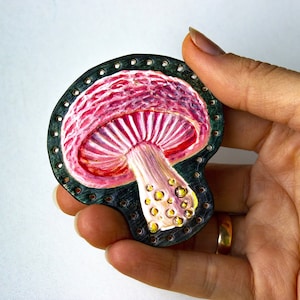 Puede incluir: Broche con forma de seta pintada a mano. El sombrero de la seta es rosa y blanco con aspecto texturizado, y el tallo es beige con manchas amarillas. El broche está delineado con un borde verde oscuro con pequeños agujeros.