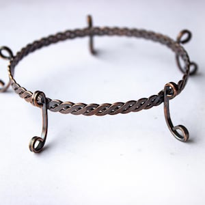 Könnte beinhalten: Ein handgefertigter, kreisförmiger Metallständer mit geflochtenem Design. Der Ständer besteht aus dunkelbraunem Metall und hat gebogene Beine und eine Schlaufe oben. Er dient wahrscheinlich zur Präsentation eines dekorativen Gegenstands. Der Durchmesser beträgt ca. 15 cm.
