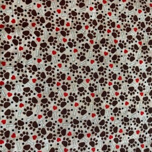 May include: A fabric pattern featuring a repeating design of brown paw prints, red hearts, white bones, and black dots on a light beige background. The pattern is evenly spaced and covers the entire surface.