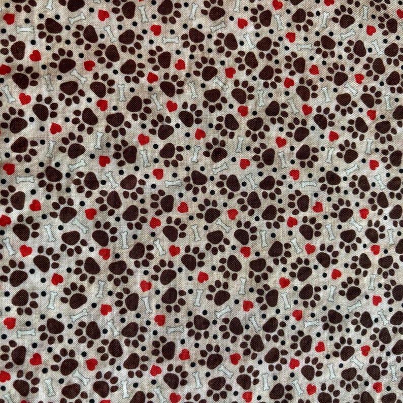 May include: A fabric pattern featuring a repeating design of brown paw prints, red hearts, white bones, and black dots on a light beige background. The pattern is evenly spaced and covers the entire surface.
