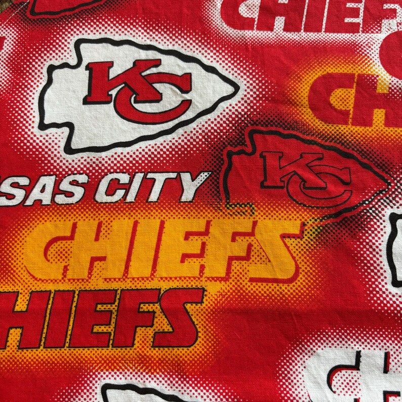 May include: Red fabric with a repeating pattern featuring the Kansas City Chiefs logo and text. The logo is a white arrowhead with a red "K" and "C". The text "KANSAS CITY CHIEFS" is also visible.