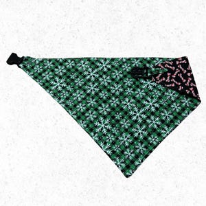 May include: Reversible dog bandana with a black buckle. One side features a green and black plaid pattern with white snowflakes. The other side has a black background with red and white candy canes.