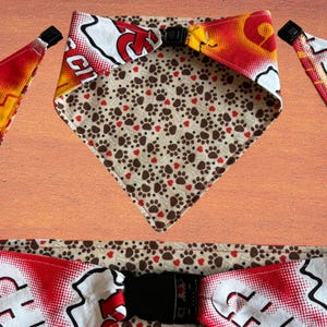 May include: Dog bandana with a reversible design. One side features a pattern of brown paw prints and red hearts on a tan background. The other side has a red, white, and yellow design with a black buckle closure. The bandana is made of fabric.