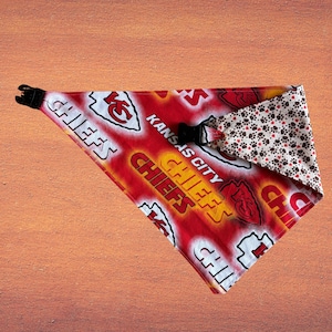 May include: Red and white Kansas City Chiefs dog bandana with the team logo and the word "CHIEFS" printed on it. The bandana has a black buckle and a reversible side with paw prints.