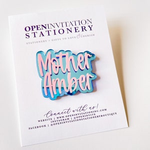May include: A blue and pink acrylic pin with the words "Mother Amber" on a white card. The pin has a reflective surface and is in a cursive font. The card has the text "OPENINVITATION STATIONERY" and "Connect with us!" printed on it.