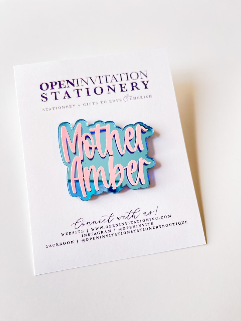 May include: A blue and pink acrylic pin with the words "Mother Amber" on a white card. The pin has a reflective surface and is in a cursive font. The card has the text "OPENINVITATION STATIONERY" and "Connect with us!" printed on it.