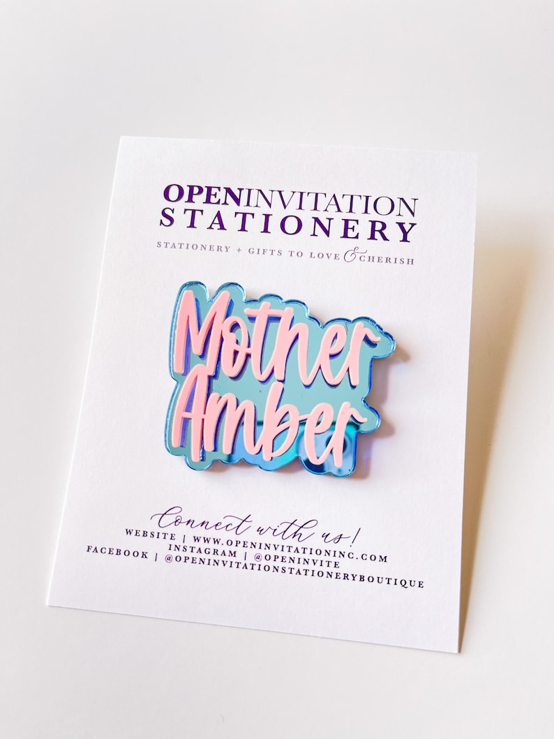 May include: A layered acrylic pin with the words "Mother Amber" in pink over a blue background. The pin is on a white card with the text "OPEN INVITATION STATIONERY" and "STATIONERY + GIFTS TO LOVE & CHERISH".