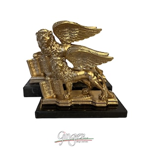 Könnte beinhalten: Zwei goldfarbene Löwenstatuen mit Flügeln, die jeweils ein Buch halten, stehen auf einem schwarzen Marmorsockel. Die Statuen haben detaillierte Mähnen und Flügel. Das Bild ist vor einem weißen Hintergrund.